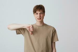 avvicinamento di triste deluso giovane uomo nel beige t camicia con bretelle su denti sembra irritato e mostrando pollici giù isolato al di sopra di bianca sfondo foto