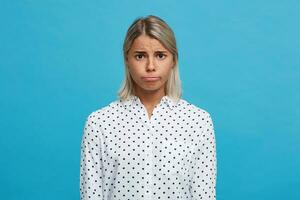 ritratto di irritato scontento bionda giovane donna indossa polka punto camicia fa triste viso e si sente depresso isolato al di sopra di blu sfondo foto