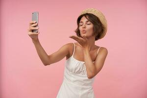 piacevole guardare giovane brunetta signora nel cannuccia cappello e bianca elegante vestito conservazione smartphone nel sollevato mano e soffiaggio aria bacio per telecamera con chiuso occhi, isolato al di sopra di rosa sfondo foto