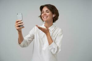 positivo bella brunetta donna con corto taglio di capelli fabbricazione foto di se stessa con sua smartphone, sorridente ampiamente per telecamera e raccolta felicemente sua palma, isolato al di sopra di bianca sfondo