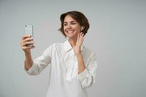 allegro bello giovane femmina con corto Marrone capelli conservazione mobile Telefono nel sollevato mano e sorridente per telecamera, indossare bianca camicia mentre in piedi al di sopra di bianca sfondo foto