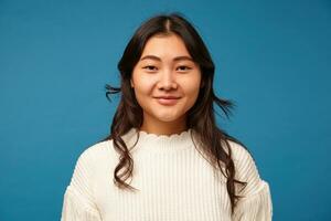 interno foto di piacevole guardare giovane buio dai capelli asiatico femmina con ondulato acconciatura sorridente positivamente a telecamera mentre in piedi contro blu sfondo