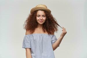 adolescenziale ragazza, contento guardare donna con Riccio Zenzero capelli. indossare a strisce spalle scoperte camicetta e cappello. giocando con filo di capelli, Sorridi. Guardando a il telecamera, isolato al di sopra di bianca sfondo foto