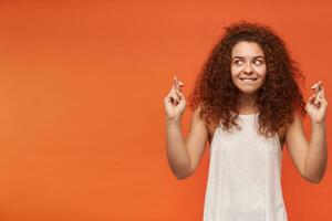 ritratto di attraente, adulto testa Rossa ragazza con Riccio capelli. indossare bianca senza spalle camicetta. mantiene dita attraversato, fabbricazione un' desiderio. Guardando per il sinistra a copia spazio, isolato al di sopra di arancia sfondo foto