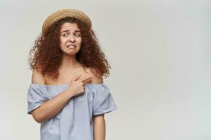 infelice guardare donna, ragazza con Riccio Zenzero capelli. indossare a strisce spalle scoperte camicetta e cappello. Guardando per il sinistra e puntamento per il giusto a copia spazio, isolato al di sopra di arancia sfondo foto