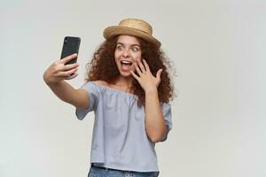 ritratto di attraente, adulto testa Rossa ragazza con Riccio capelli. indossare a strisce spalle scoperte camicetta e cappello. assunzione un' autoscatto su un' smartphone e toccante sua viso. In piedi isolato al di sopra di bianca sfondo foto