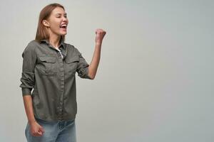 giovane signora, bella donna con Marrone lungo capelli. indossare grigio camicia e jeans. mantenere cazzotto sollevato, trasformato e Guardando per il giusto a copia spazio, mostrando sua eccitazione, isolato al di sopra di grigio sfondo foto