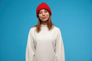 rilassato guardare donna, bellissimo ragazza con brunetta capelli. indossare bianca maglione e rosso cappello. persone e emozione concetto. Guardando a il telecamera e Sorridi, isolato al di sopra di blu sfondo foto