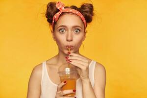 ritratto di attraente, bello ragazza con Due panini e grande occhi. indossare bianca serbatoio superiore e rosso stravinto cerchietto per capelli. avendo un' sorso di salutare succo. Guardando a il telecamera isolato al di sopra di giallo sfondo foto