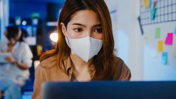 felice donna d'affari asiatica che indossa una maschera medica per il distanziamento sociale in una nuova situazione normale per la prevenzione dei virus mentre si utilizza il laptop al lavoro durante la notte in ufficio. vita e lavoro dopo il coronavirus.