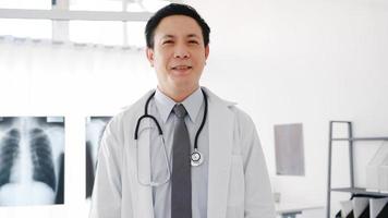 giovane medico maschio asiatico in uniforme medica bianca con stetoscopio che guarda l'obbiettivo, sorriso e braccia incrociate durante la videoconferenza con il paziente in ospedale. concetto di consulenza e terapia. foto