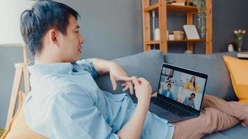 il giovane uomo d'affari asiatico che utilizza il laptop parla con i colleghi del piano in una riunione di videochiamata mentre lavora da casa in soggiorno. autoisolamento, distanziamento sociale, quarantena per la prevenzione del virus corona.