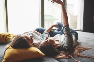 due sorelle gemelle sorridenti carine che tengono smartphone e fanno selfie. le ragazze si sdraiano sul divano in posa e gioia foto