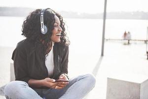 ritratto di una giovane e bella ragazza afroamericana seduta sulla spiaggia o sul lago e ascoltando musica nelle sue cuffie