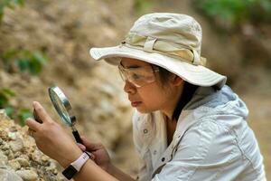 femmina geologo utilizzando ingrandimento bicchiere per esaminare e analizzare roccia, suolo, sabbia nel natura. archeologi Esplorare il campo. ambientale e ecologia ricerca. foto