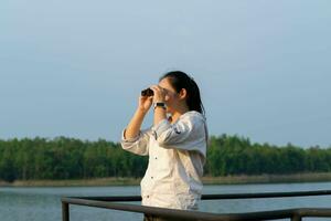 giovane femmina esploratore con binocolo esplorando natura o Guardando uccelli all'aperto. giovane donna guardare attraverso binocolo a uccelli su il serbatoio. osservazione uccelli foto