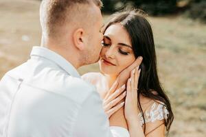 un' elegante coppia di europeo Novelli sposi. un' sorridente sposa nel un' bianca vestito abbracci il sposo a partire dal dietro. il sposo è vestito nel un' bianca camicia, senza un' legare. all'aperto nozze nel il montagne foto