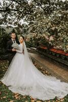 classico lunghezza intera ritratto di il sposa e sposo nel il parco tra verdura e quercia foglie, elegante vestito con un' lungo treno. gratuito spazio foto