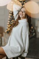 un' bellissimo e elegante giovane ragazza nel un' bianca maglione pose nel il Natale luci su il sfondo di il Natale albero foto