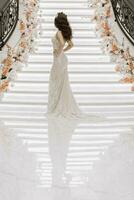 bellissimo ragazza sposa, con un' bianca nozze vestito e un' lussuoso corona su sua testa, lungo Marrone capelli, vicino il le scale decorato con fiori. verticale foto