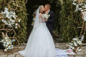 un' giovane nozze coppia a un' nozze pittura cerimonia. il sposa e sposo bacio contro il sfondo di verde alberi e un arco decorato con fiori foto