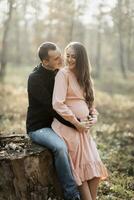 un' uomo e il suo incinta moglie abbraccio e bacio nel il foresta a tramonto. contento famiglia concetto. foto