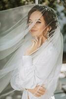 ritratto di un' sognante sposa in piedi di un' letto nel un' giardino sotto un' velo. il sposa è vestito nel un' bianca accappatoio e un' tiara di fresco fiori su sua testa foto
