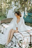 il mattina di il sposa su il letto nel il giardino sotto il albero. il sposa è indossare un' bianca vestito e un' tiara di fresco fiori su sua testa foto