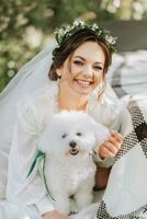 giovane elegante sposa nel un' bianca vestito con un' tiara di fresco fiori su sua testa in posa nel il giardino con sua barboncino cane foto