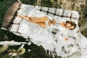 ritratto di un' sognante sposa dire bugie su un' letto nel un' giardino sotto un' albero. il sposa è indossare un' bianca vestito e un' tiara di fresco fiori su sua testa foto