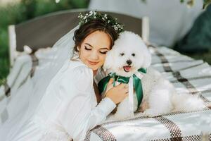 giovane elegante sposa nel un' bianca vestito con un' tiara di fresco fiori su sua testa in posa nel il giardino con sua barboncino cane foto