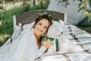 giovane elegante sposa nel un' bianca vestito con un' tiara di fresco fiori su sua testa in posa nel il giardino con sua barboncino cane foto