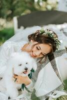 giovane elegante sposa nel un' bianca vestito con un' tiara di fresco fiori su sua testa in posa nel il giardino con sua barboncino cane foto