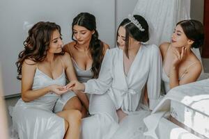 giovane damigelle d'onore nel bianca seta vestiti su un' letto nel il della sposa camera. bellissimo donne festeggiare addio al nubilato festa seduta su letto. foto