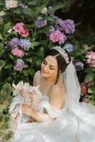 giovane bellissimo sposa nel nozze vestito con Aperto le spalle e corona su sua testa seduta vicino ortensia fiori, moda foto prese sotto duro luce del sole