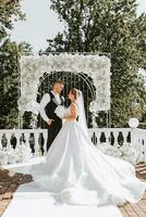 elegante sposo nel bianca camicia e nero veste e carino bionda sposa nel bianca vestito nel parco vicino elegante bianca rosa arco. nozze ritratto di Novelli sposi. foto