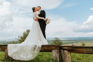 elegante sposa e sposo abbraccio e bacio contro il sfondo di estate montagne. il concetto di un' rustico nozze nel il montagne, contento boemo Novelli sposi. foto