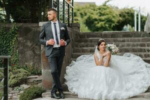 giovane bellissimo sposa con blu occhi nel un' nozze vestito con un' mazzo di fiori nel sua mani, seduta su il pietra passi, sua marito Il prossimo per suo. foto