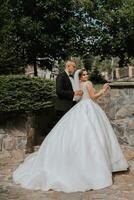 nozze ritratto. il sposa nel un elegante vestito sta nel davanti di il sposo nel un' classico completo da uomo, contro il sfondo di verde alberi. dolce tocco. estate nozze. un' camminare nel natura foto