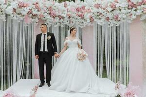 attraente sposa e sposo a il cerimonia su loro nozze giorno con un arco fatto di rosa e bianca fiori. bellissimo Novelli sposi, un' giovane donna nel un' bianca vestito con un' lungo treno, uomini nel un' nero completo da uomo. foto