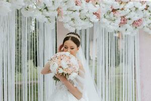 attraente sposa a il cerimonia su sua nozze giorno con un arco fatto di rosa e bianca fiori. bellissimo Novelli sposi, un' giovane donna nel un' bianca vestito con un' lungo treno, uomini nel un' nero completo da uomo. foto