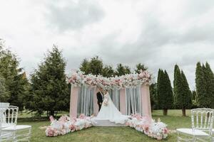 attraente sposa e sposo a il cerimonia su loro nozze giorno con un arco fatto di rosa e bianca fiori. bellissimo Novelli sposi, un' giovane donna nel un' bianca vestito con un' lungo treno, uomini nel un' nero completo da uomo. foto