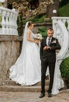 ritratto di un' giovane sposa nel un' bianca vestito e un' brunetta sposo nel un' completo da uomo su il le scale vicino il vecchio città parco. il uomo nel il primo piano. un' bellissimo e romantico nozze, un' contento coppia foto