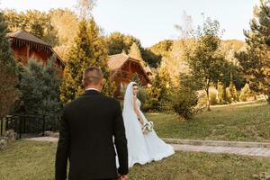 un' elegante sposo nel un' nero completo da uomo e un' carino sposa nel un' bianca vestito con un' lungo velo siamo abbracciare e a piedi vicino verde alto alberi. nozze ritratto di sorridente e contento Novelli sposi. foto