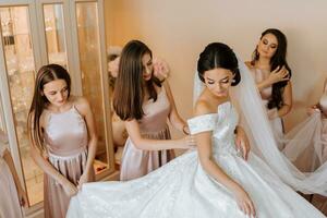 elegante bellissimo damigelle d'onore nel accoppiamento seta vestiti porzione bellissima brunetta sposa nel bianca vestito ottenere pronto per nozze, mattina preparativi, donna mettendo su vestito foto