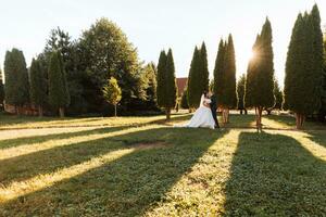 contento sposa e sposo siamo a piedi nel il parco dopo il nozze cerimonia. lungo velo. elegante bianca vestito con Aperto le spalle. un' mazzo di orchidee. durante tramonto foto