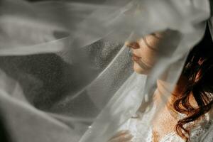 bellissimo sposa con lungo buio Riccio capelli nel un' lungo bianca accappatoio nel il mattina. il sposa è preparazione per il nozze. professionale trucco e capelli. un' corona su il testa e un' lungo velo di il sposa foto