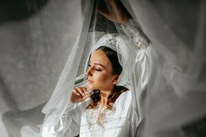 bellissimo sposa con lungo buio Riccio capelli nel un' lungo bianca accappatoio nel il mattina. il sposa è preparazione per il nozze. professionale trucco e capelli. un' corona su il testa e un' lungo velo di il sposa foto