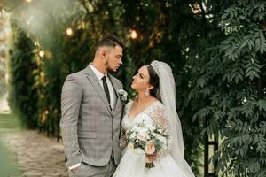 giovane nozze coppia godendo romantico momenti all'aperto su un' estate prato. un' sposa con un' corona su sua testa e un' lungo velo. sposo nel un' classico grigio completo da uomo foto