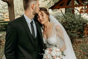 bellissima elegante lussuoso sposa con velo soffiaggio nel il vento. e un' elegante sposo bacio all'aperto vicino alto autunno alberi foto